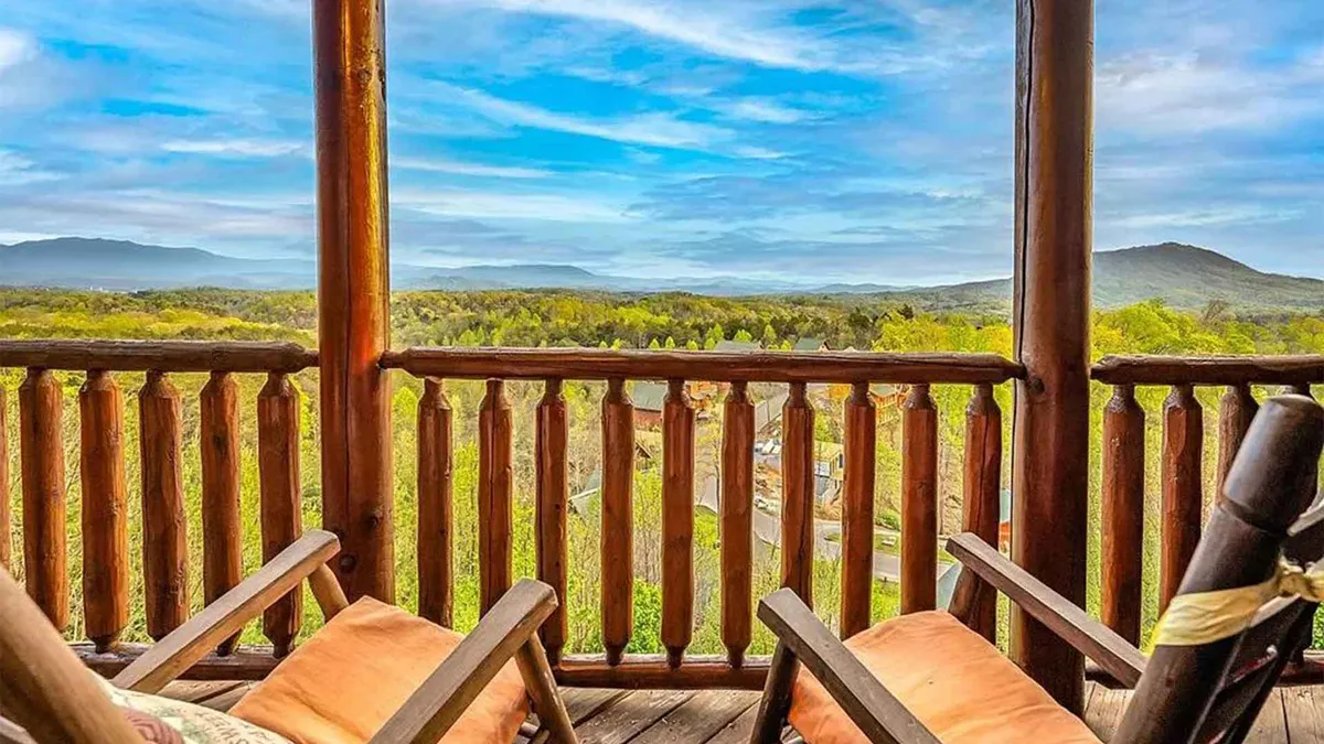 A stunning exterior shot from the cabin's balcony, featuring classic wooden railings and comfortable rocking chairs. The view stretches across the lush green landscape to the distant Smoky Mountains beneath a vibrant blue sky, perfectly capturing the tranquil beauty of the setting.