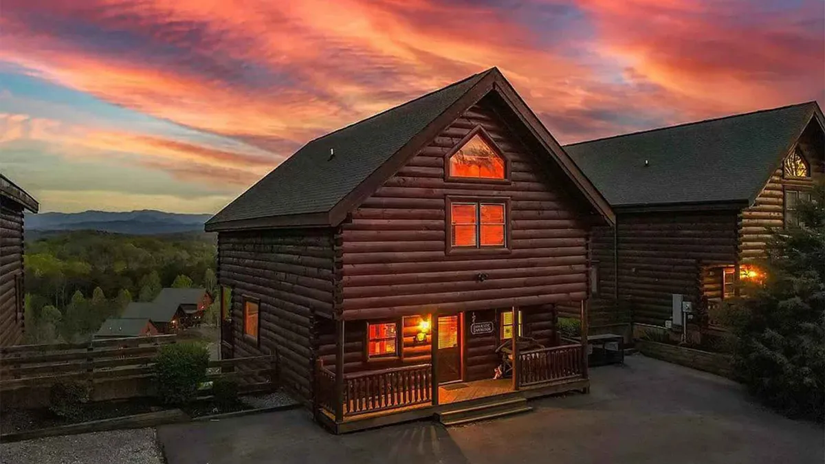 The image showcases a stunning log cabin basking in the golden glow of sunset, with warm light streaming from its windows. The expansive porch invites relaxation while the panoramic Smoky Mountain views stretch across the horizon. Guests can be seen gathering around a cozy fire pit, perfectly capturing the warmth and togetherness ideal for a mountain getaway.