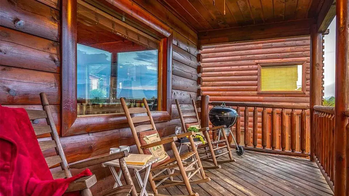The image shows a cozy porch with rocking chairs, a grill, a wooden railing, and a large window. The sunset light highlights a relaxed atmosphere perfect for enjoying nature, barbecuing, or sharing quiet moments together.