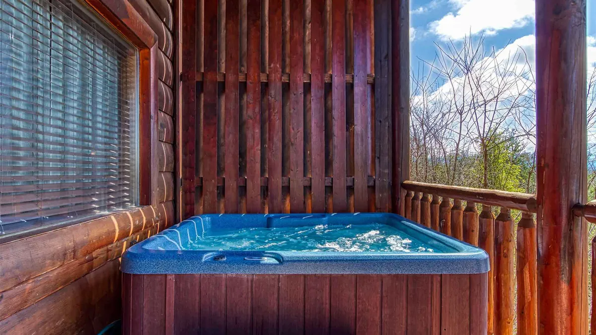 A bubbling hot tub nestled on the deck, framed by a wooden railing and adjacent window, offers a peaceful retreat with partial views of the surrounding landscape. The inviting water and natural wood details evoke a sense of secluded relaxation.