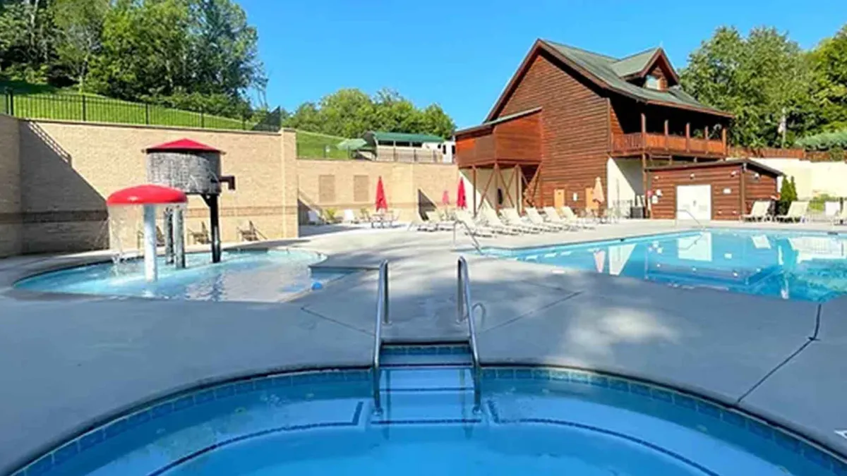 A relaxed and cheerful resort pool scene, featuring a swimming pool, hot tub, red umbrellas, and a nearby wooden building. Families and children are depicted enjoying the water, sunbathing, and gathering for poolside fun, which enhances the sense of community and shared enjoyment of amenities.