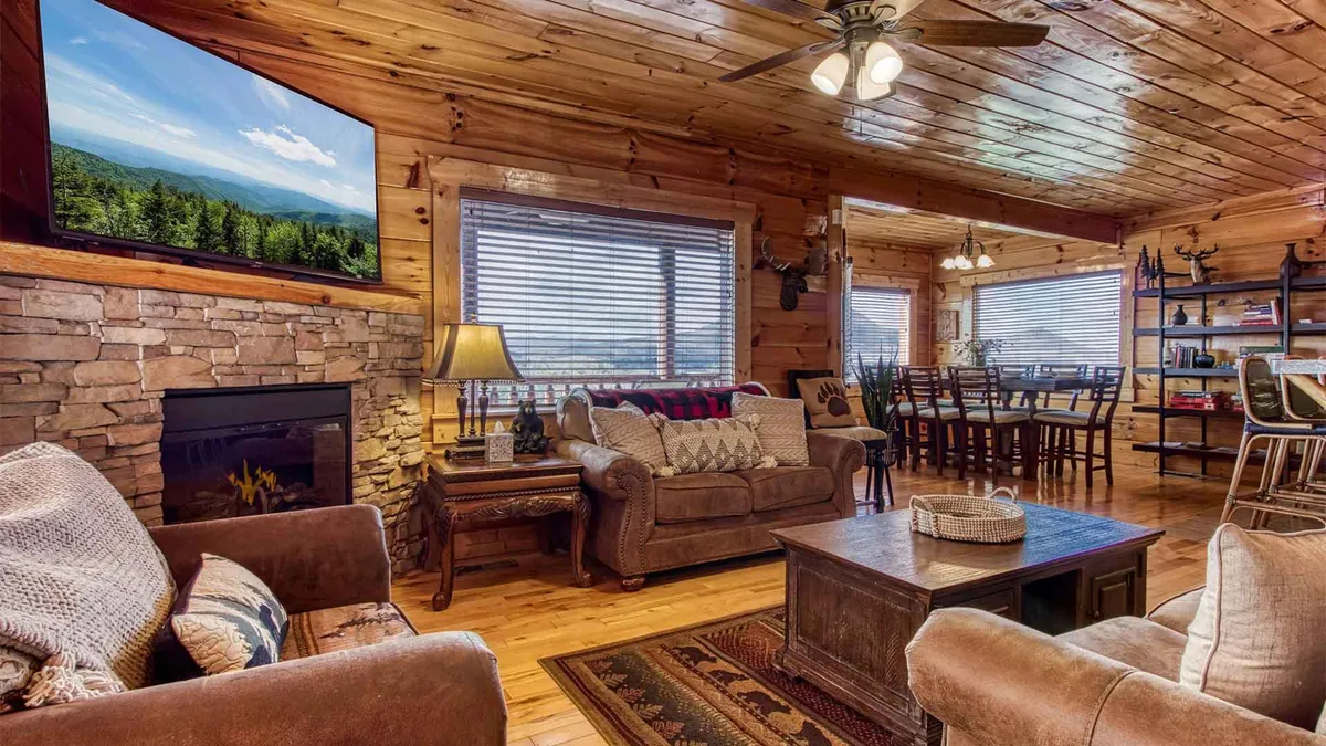 A spacious, inviting living room featuring a stone fireplace, large windows framing breathtaking mountain views, a cozy arrangement of comfortable sofas, and a wooden dining table in the background. The room is bathed in warm, natural light, creating a perfect setting for family gatherings and shared moments.