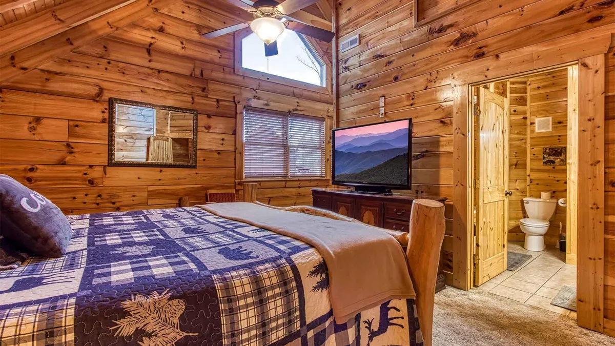 A bedroom with a wood-paneled interior, a bed dressed in a patterned quilt, a television, and a window that frames mountain views. The room feels both welcoming and connected to the natural surroundings.