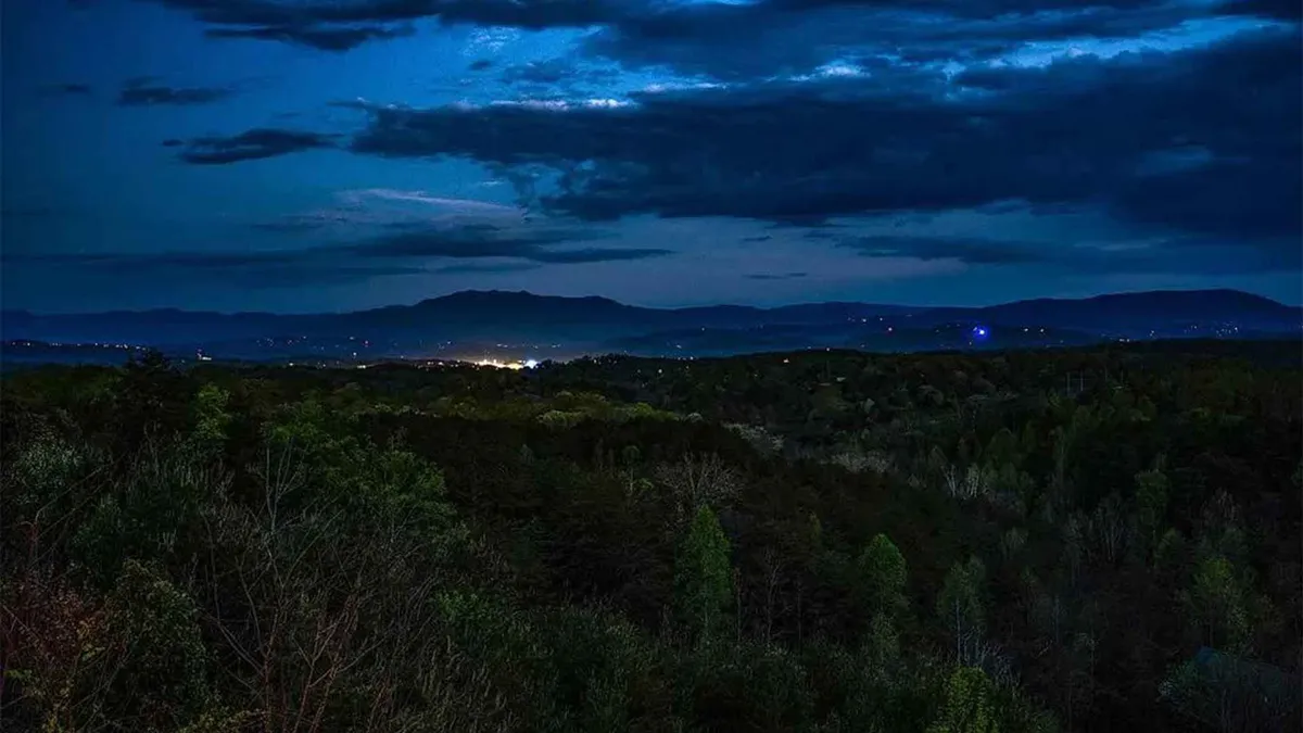 A twilight view looking out over distant mountains, with the valley below softly glowing from scattered lights. Dense forest surrounds the valley, evoking a sense of peaceful seclusion and connection to nature. The expansive night sky sets the stage for stargazing and tranquil moments.